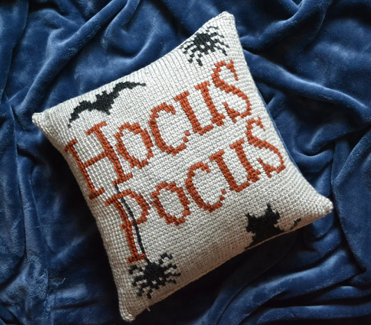 A tunisian crochet pillow cover that reads "Hocus Pocus" featuring two spiders, a bat, and a cat