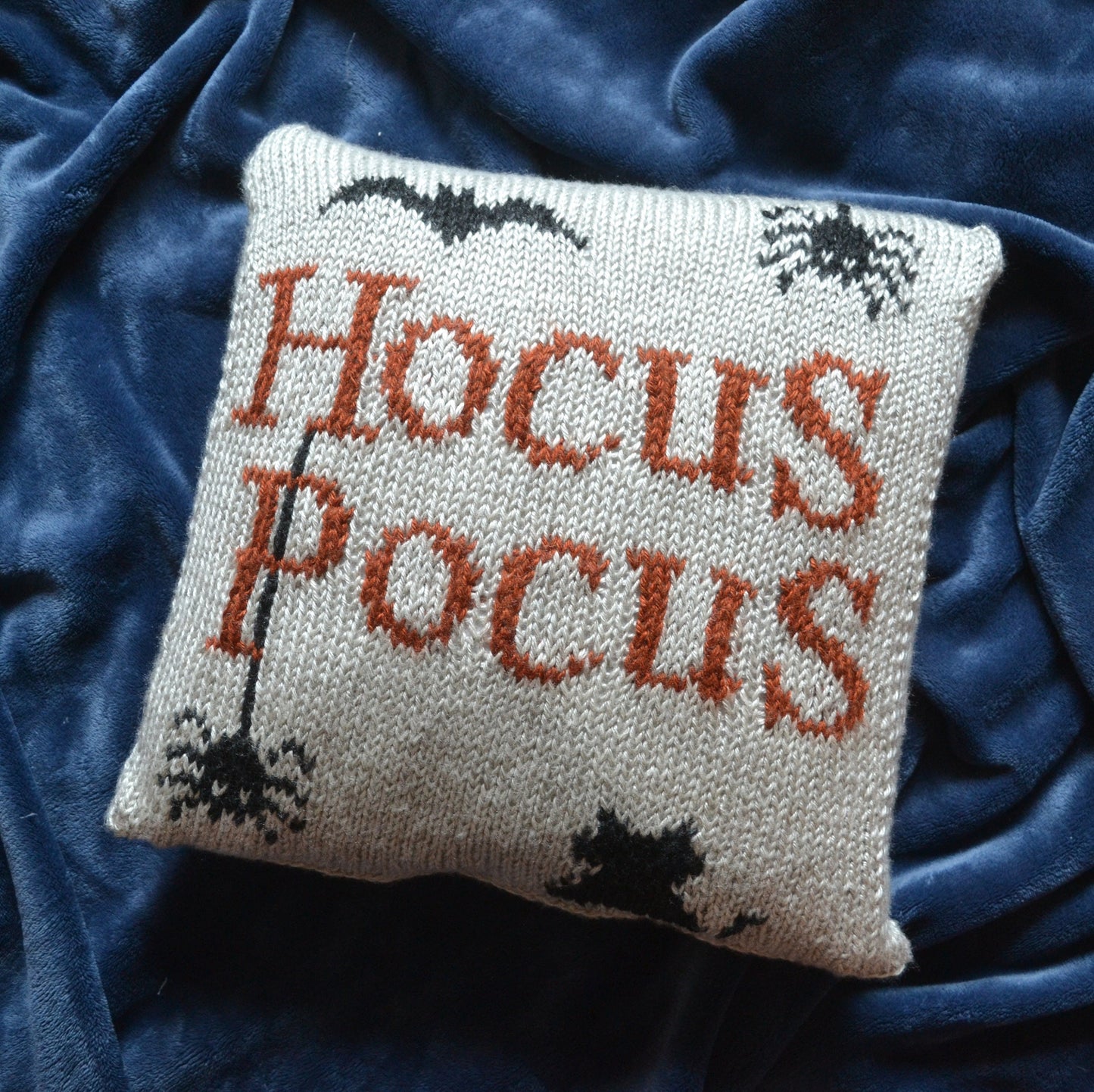 A knit pillow cover that reads "Hocus Pocus" featuring two spiders, a bat, and a cat